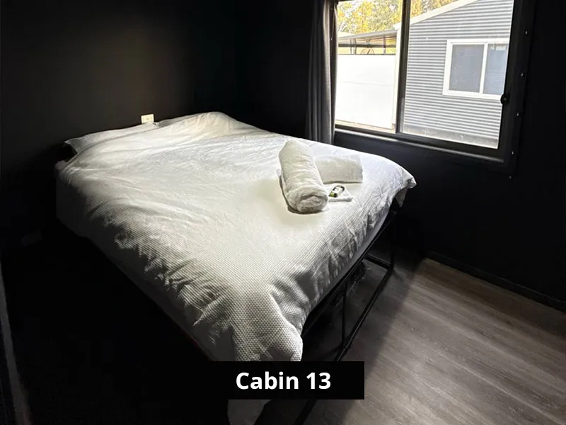 Modern dark-painted cabin bedroom with white double bed, fresh towel, large window, and wood-look flooring.