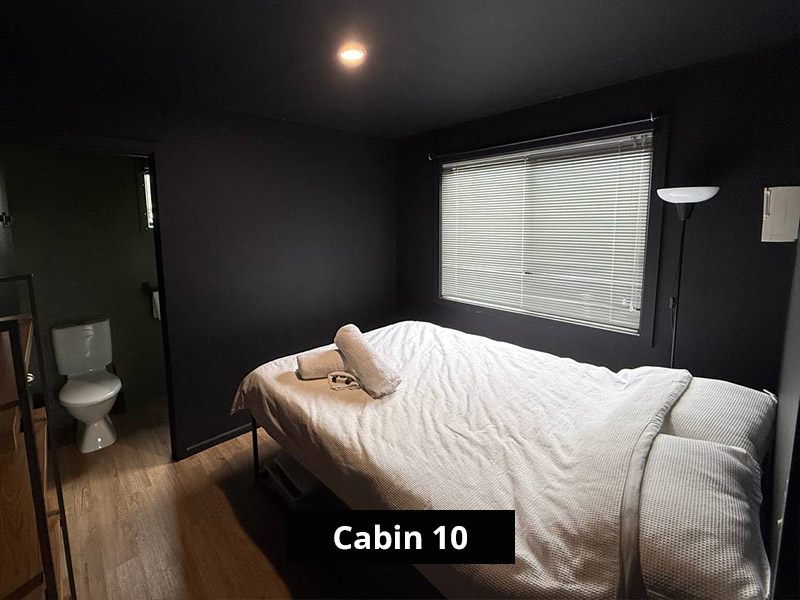 Modern dark-painted cabin bedroom with white double bed, fresh towel, large window, and wood-look flooring.